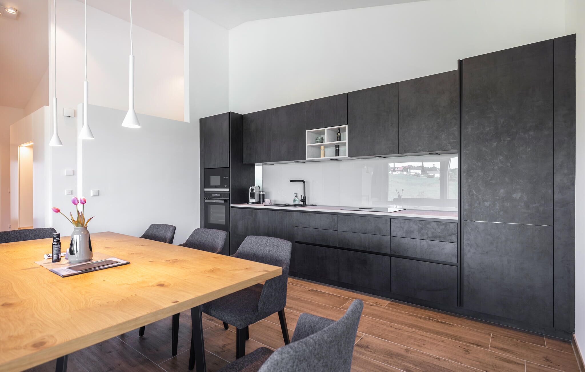 Une cuisine moderne avec des meubles de couleur sombre, une table à manger en bois et des chaises recouvertes d’un tissu gris.