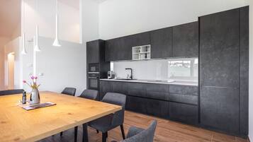 Une cuisine moderne avec des meubles de couleur sombre, une table à manger en bois et des chaises recouvertes d’un tissu gris.