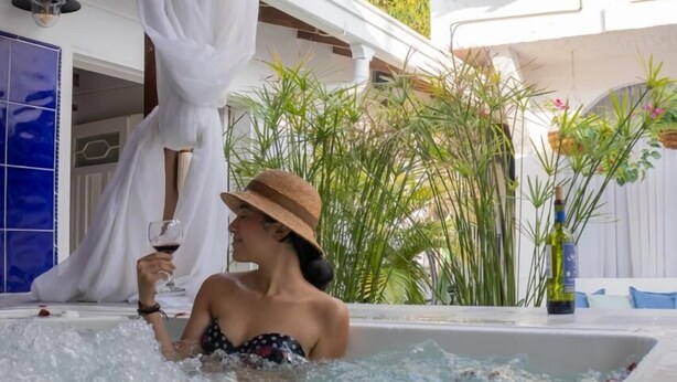 A person in a hot tub with a bottle of wine, surrounded by greenery and a white building with a balcony. - Selva Maria Hotel Boutique (Santa Fe de Antioquia)