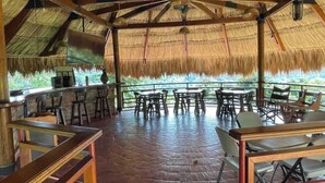 A covered outdoor dining area with thatched roof, wooden tables, and chairs.