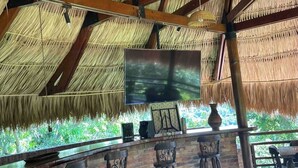 A bar area with a thatched roof, wooden bar stools, a flat-screen TV, and a view of greenery outside.
