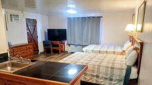 A compact hotel room with a kitchenette, a bed with a checkered duvet, a television, and a bedside lamp.