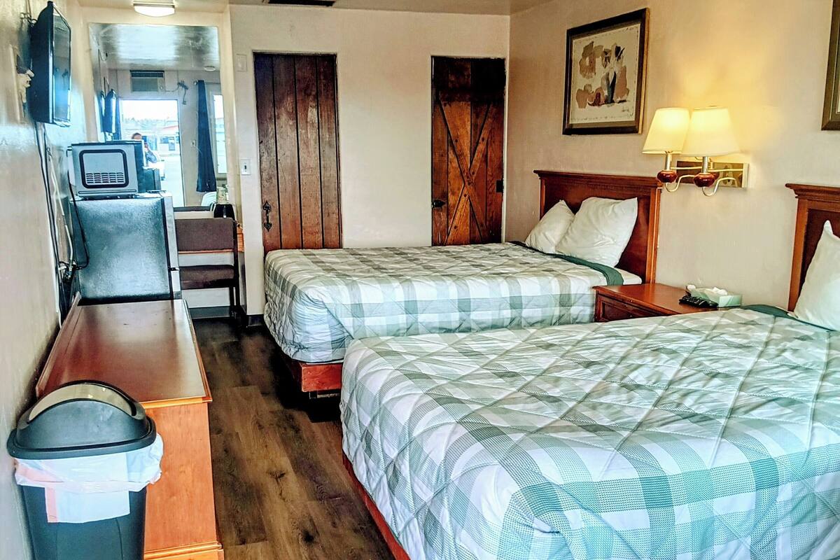 A hotel room with two beds, a desk, a trash bin, a television, and a painting on the wall.