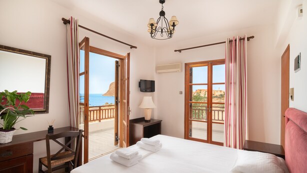 A hotel room with a bed, a desk, a chair, a TV, and a balcony with curtains. - Alta Vista Studios (Monemvasia)