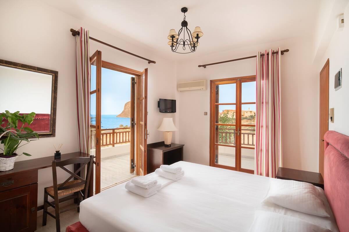 A hotel room with a bed, a desk, a chair, a TV, and a balcony with curtains.