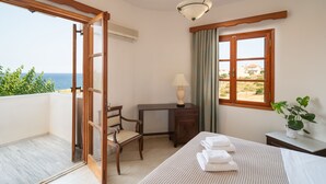 A hotel room with a bed, a desk, a chair, a window with a view of the sea, and an air conditioning unit. - Alta Vista Studios (Monemvasia)