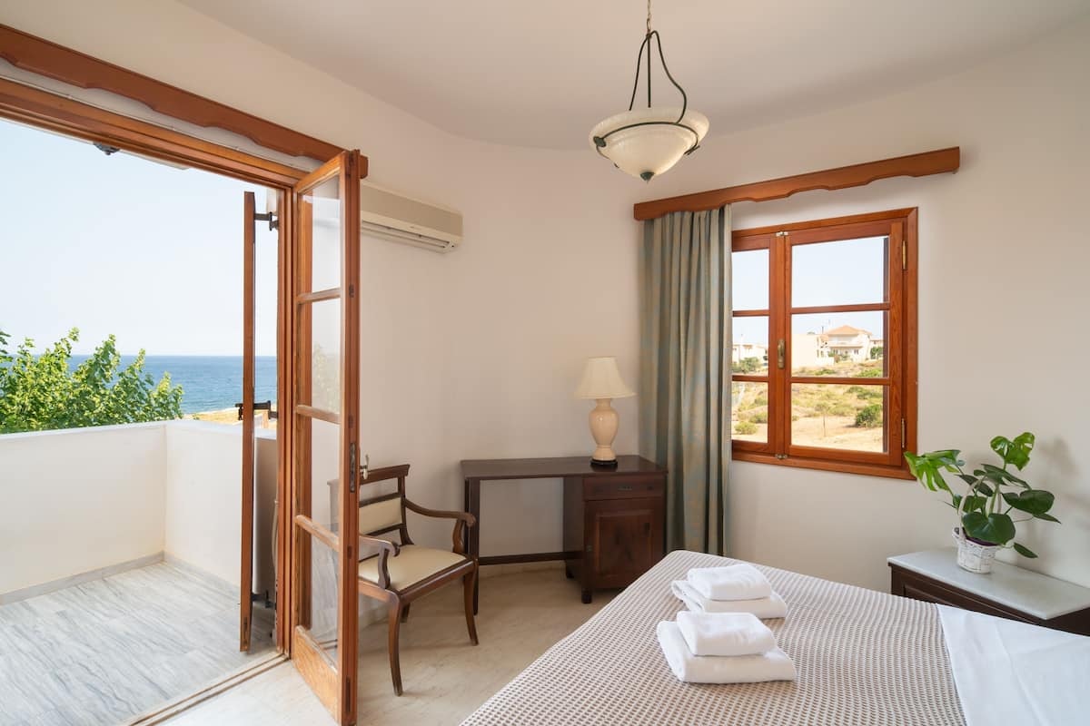 A hotel room with a bed, a desk, a chair, a window with a view of the sea, and an air conditioning unit.