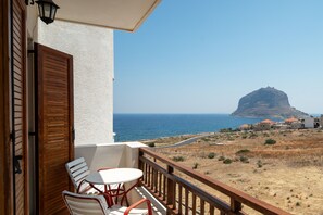 A balcony with a table and chairs, overlooking a coastal landscape with a prominent rock formation and buildings in the distance. - Alta Vista Studios (Monemvasia)