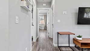 Ein Flur mit Holzboden, weißen Wänden und einem an der Wand befestigten Fernseher. Auf der rechten Seite steht ein kleiner Tisch mit einer Pflanze.