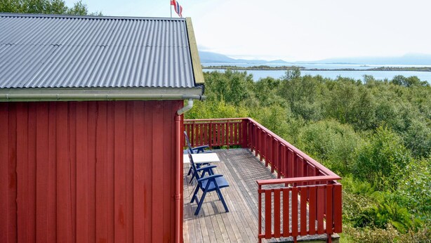 A red building with a balcony overlooking a forested area and a body of water.