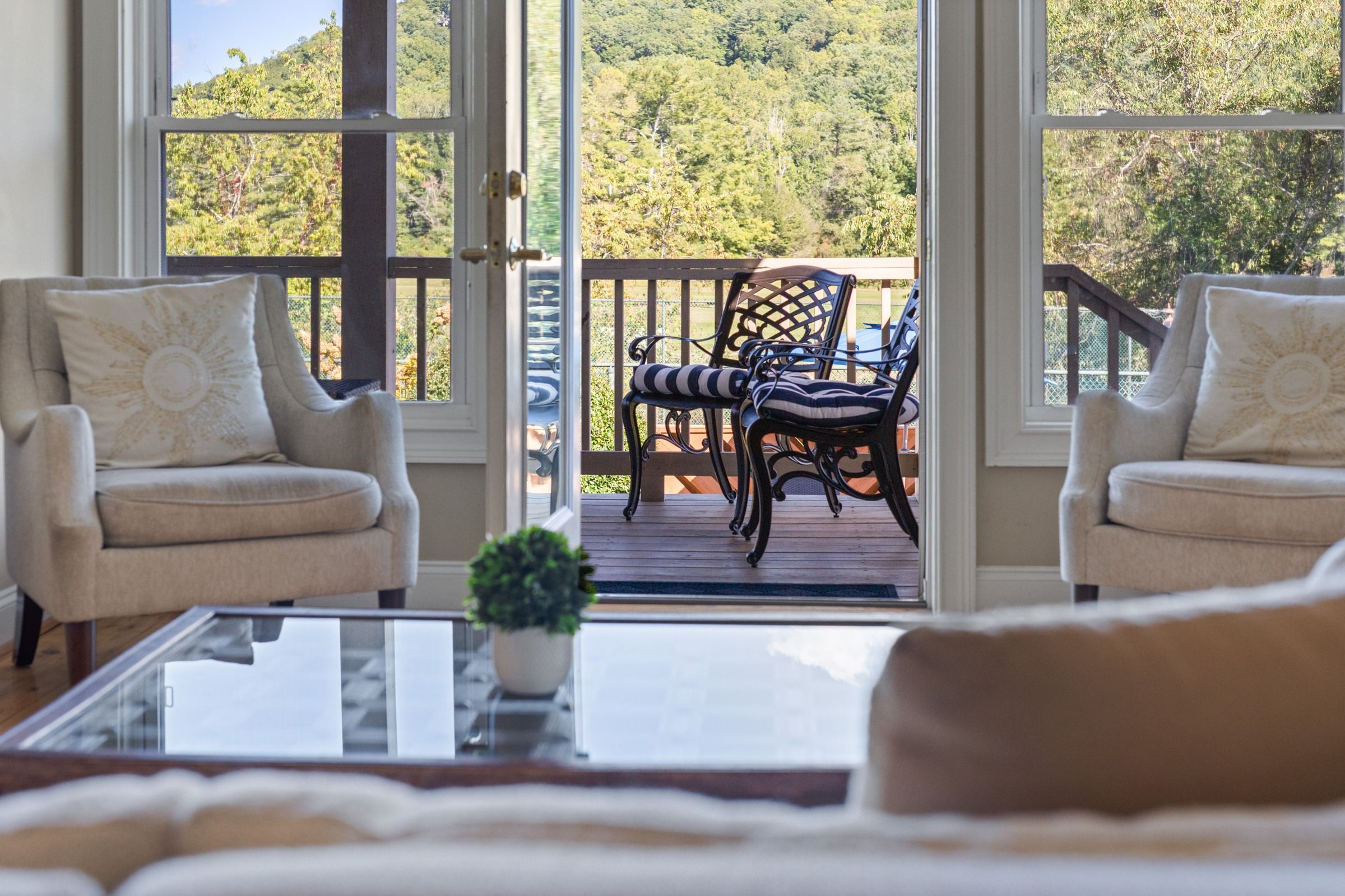 A room with two chairs facing a wooden deck with a view of a forested hill.