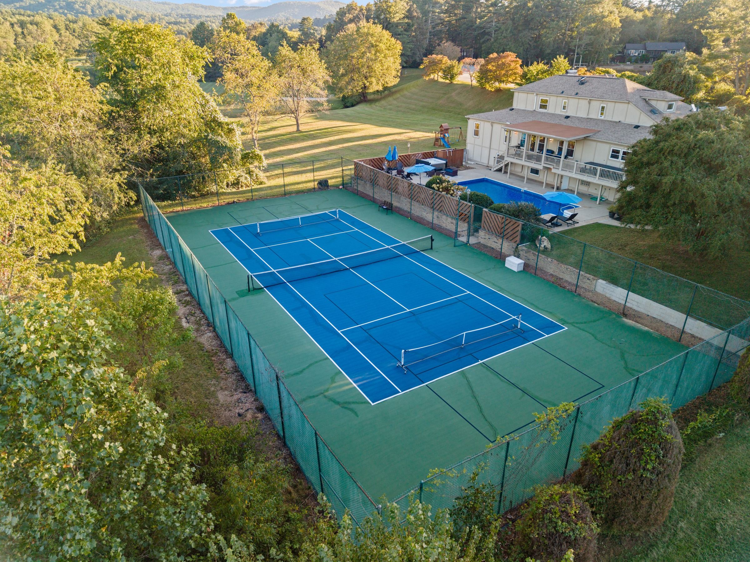 A tennis court surrounded by a green fence, with a house and trees in the background.