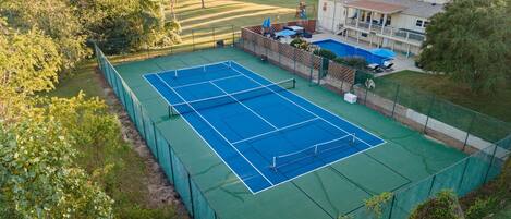 A tennis court surrounded by a green fence, with a house and trees in the background.