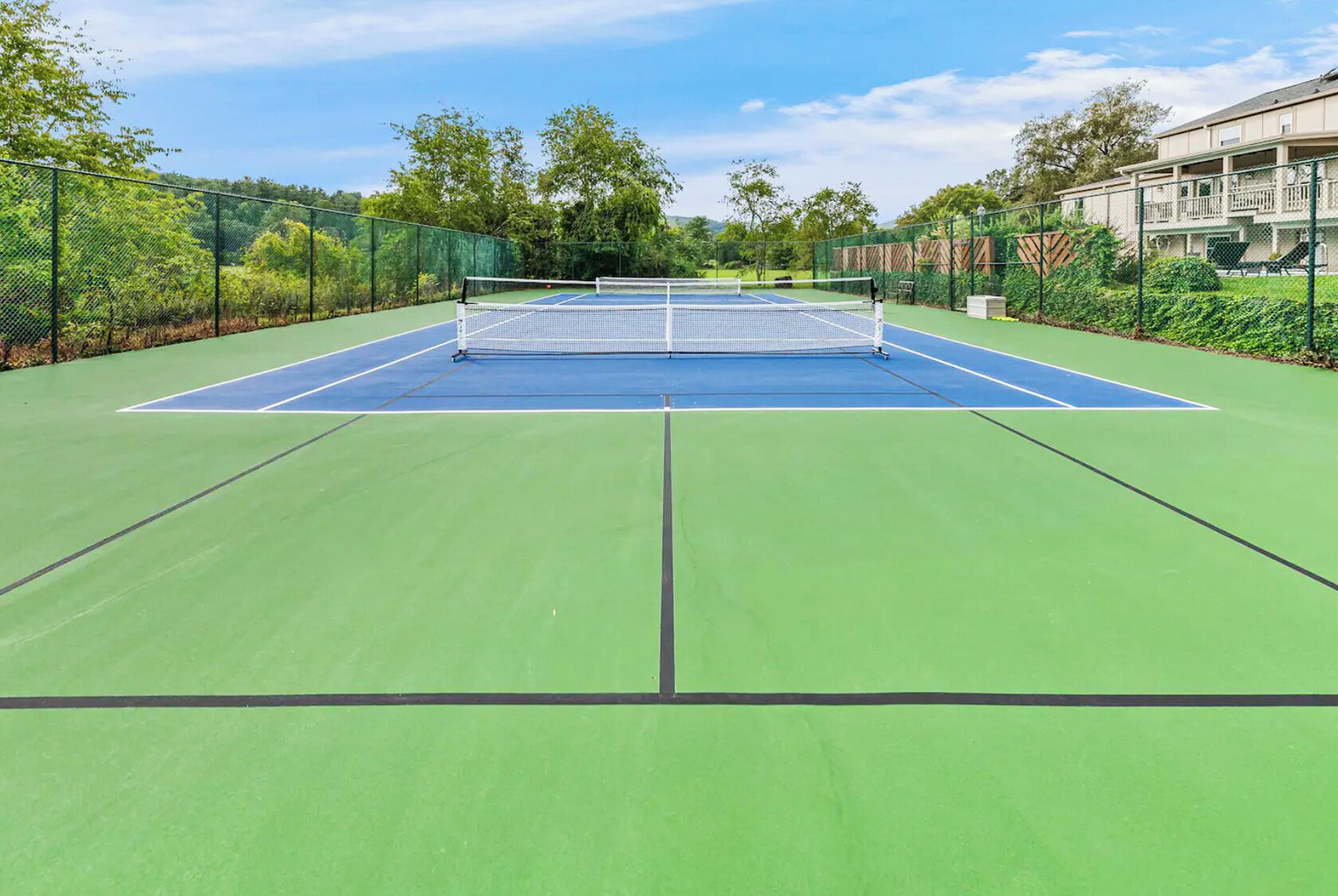 An outdoor tennis court with a net, surrounded by greenery and a building in the background.