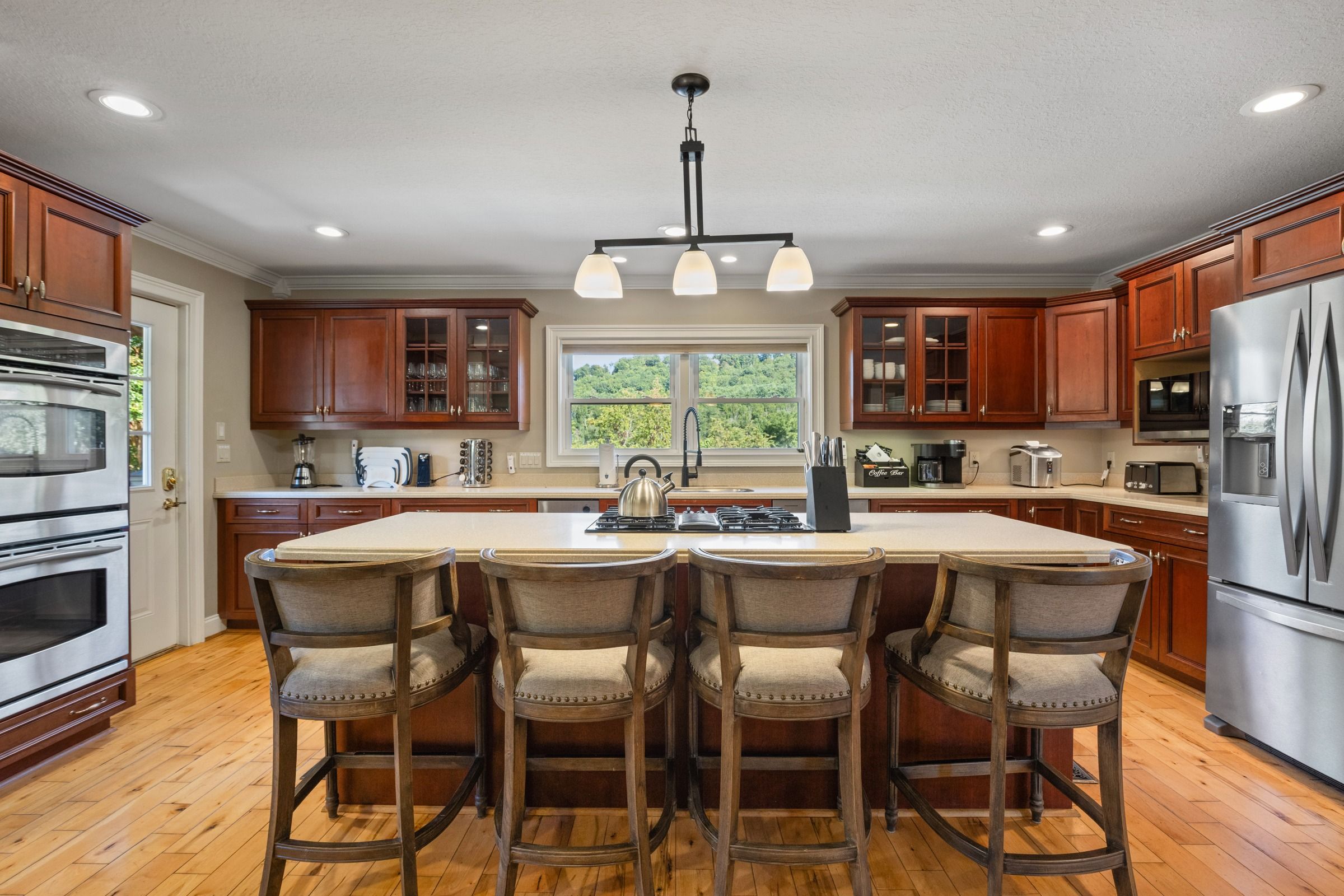 A kitchen with wooden cabinets, a stainless steel refrigerator, a large island with bar stools, and a window with a view of greenery.