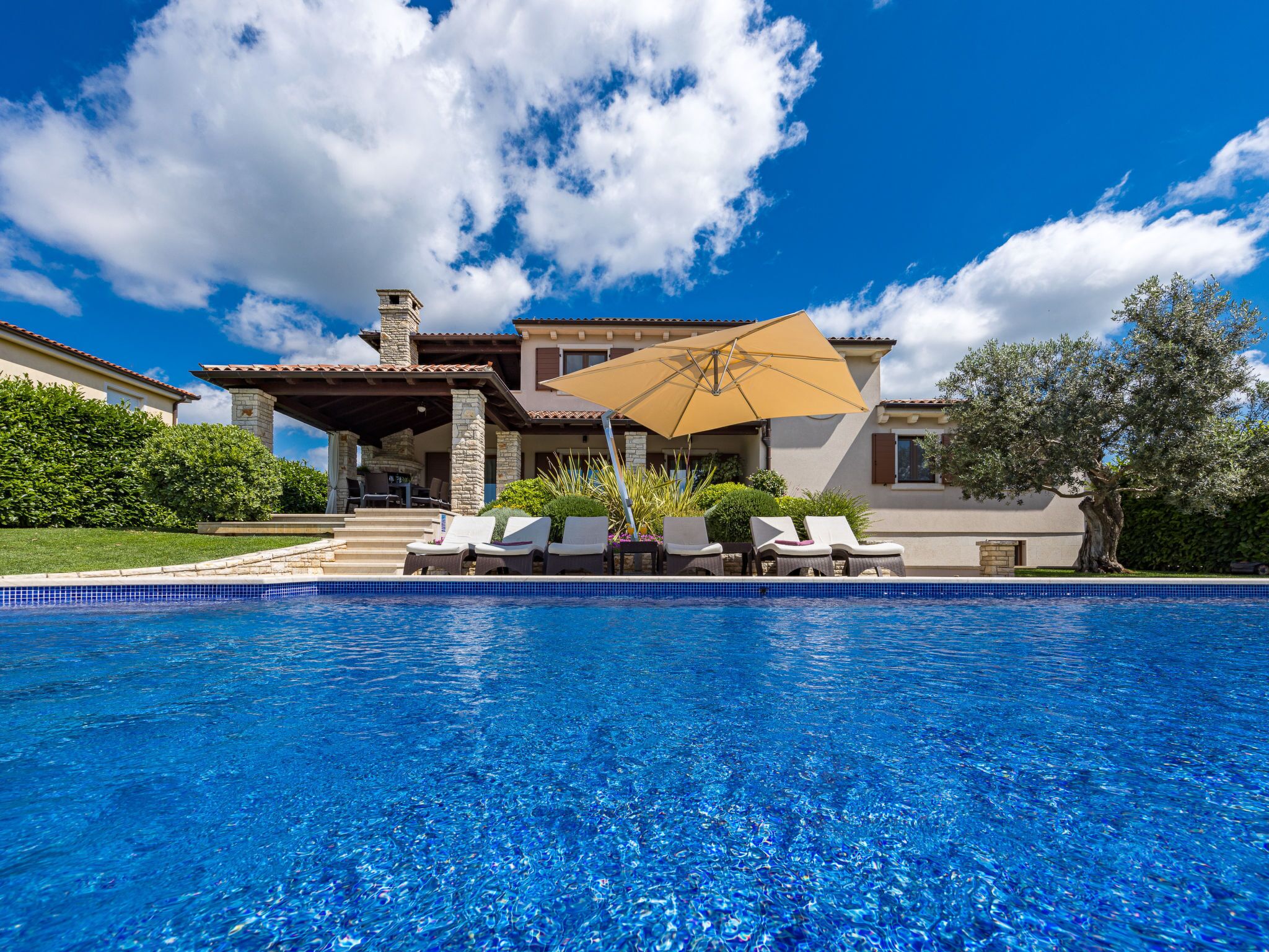 Ein Pool mit Liegestühlen, eine überdachte Terrasse und ein Haus mit Naturstein-Fassade.