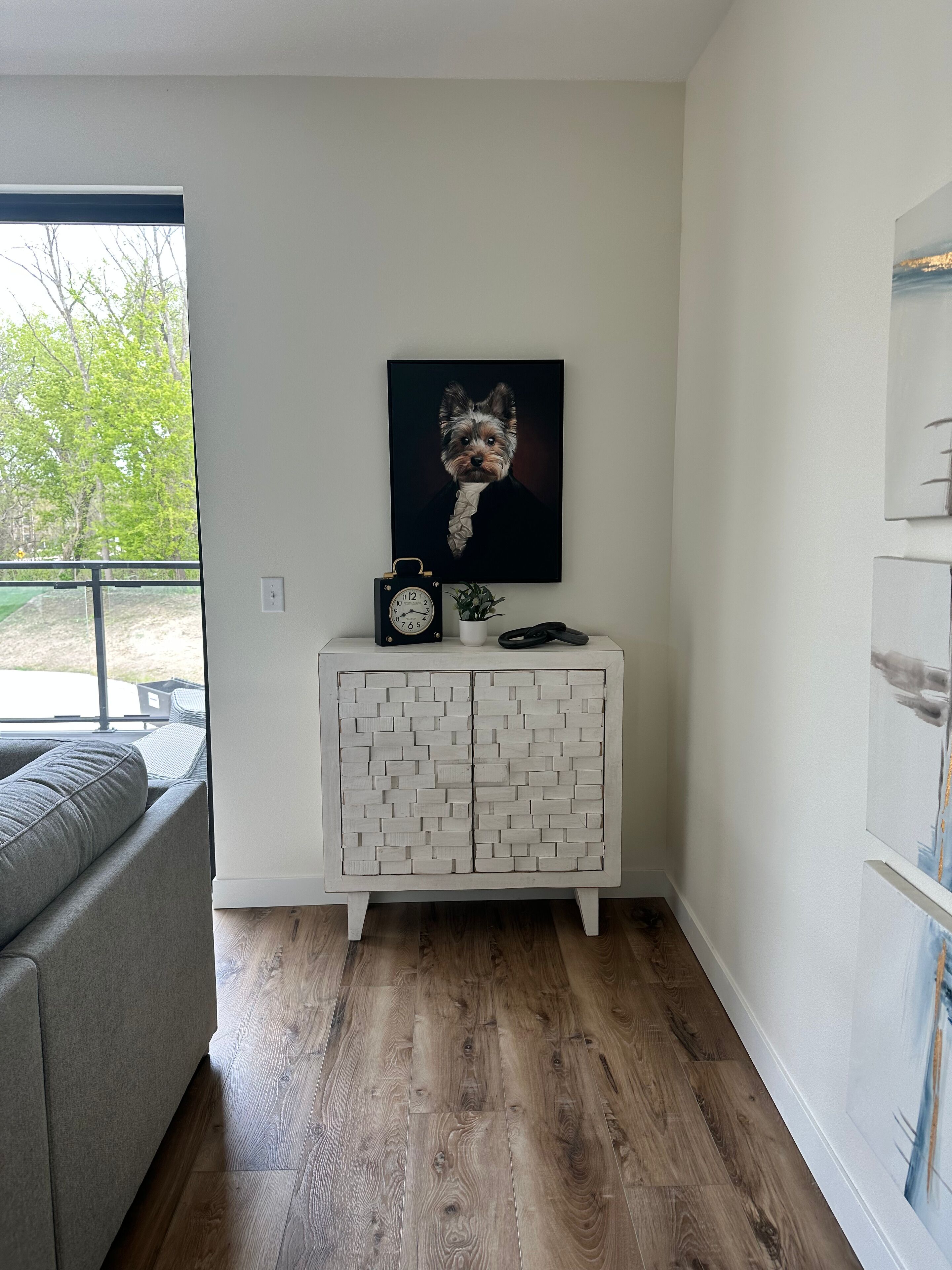 Una sala moderna con un sofá gris, un mueble blanco con diseño de ladrillo, un cuadro con la imagen de un perro y un reloj.