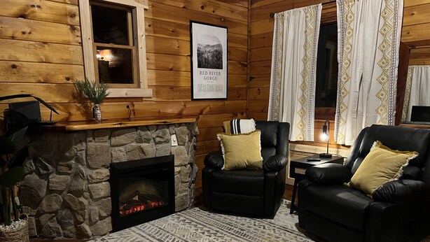 A cozy cabin interior with a stone fireplace, two leather armchairs, a patterned rug, and a framed picture of a gorge.