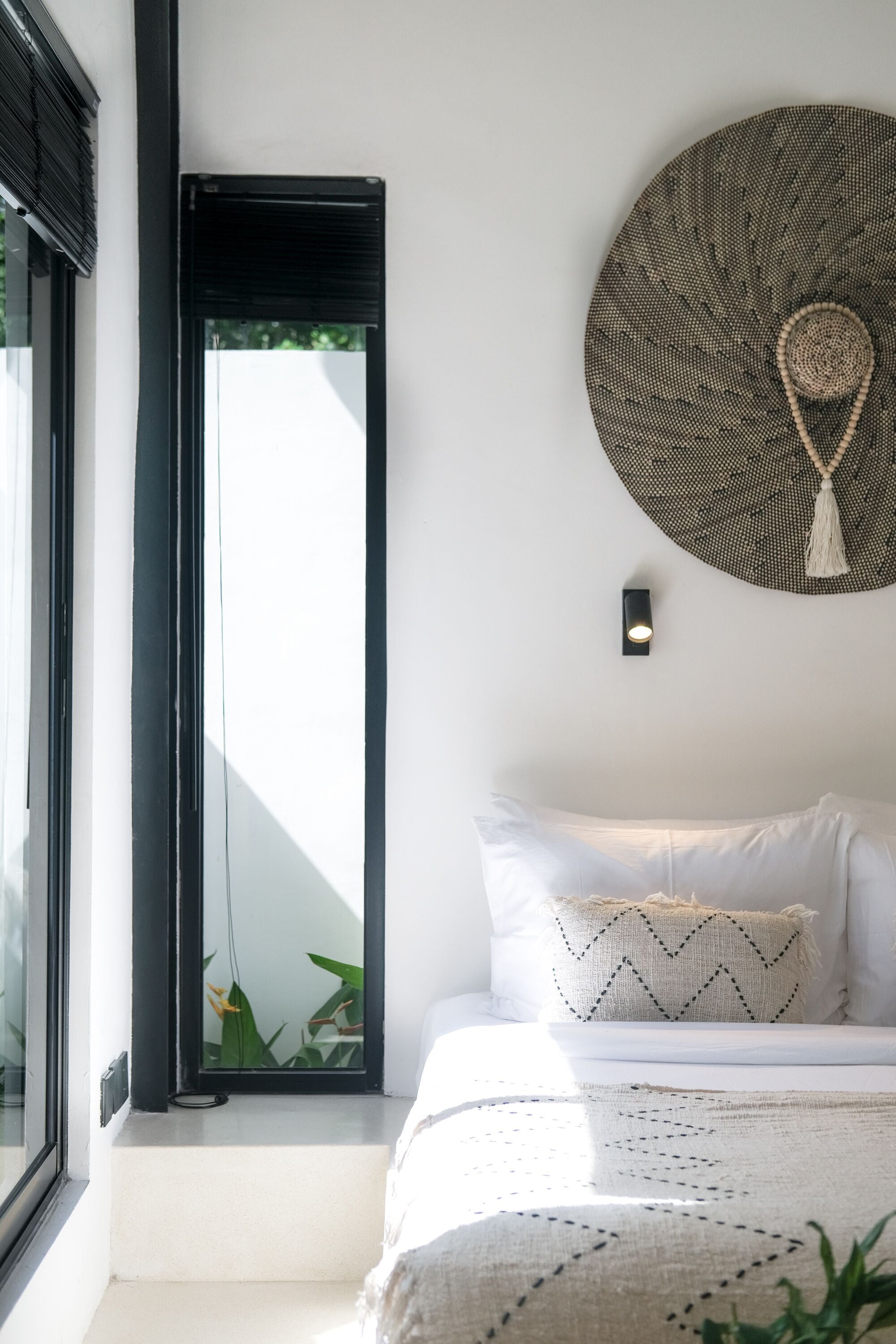 A bedroom with a bed, a window, a wall-mounted light, and a decorative wall hanging.
