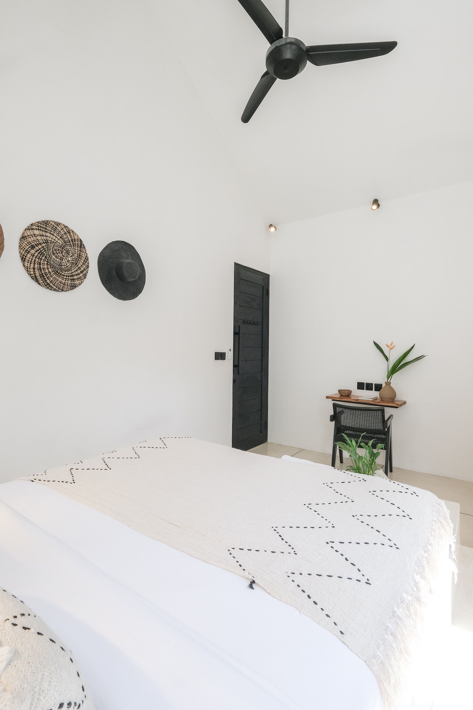 A minimalist bedroom with a bed, a ceiling fan, a door, and a small table with a plant.