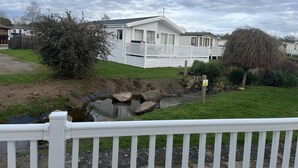 A row of white mobile homes with a small pond and a wooden fence in the foreground. - 3 bedroom stunning lodge Manor Park linen Inc (Hunstanton)