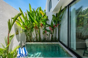 Un petit espace piscine agrémenté d’une porte en verre, entouré de plantes tropicales et bordé d’un mur blanc.