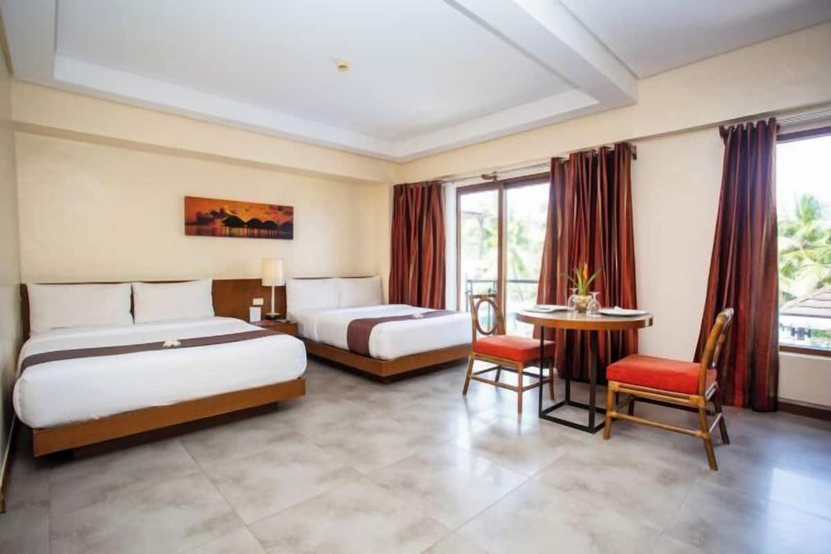 A hotel room with two beds, a small table with a chair, and a window with curtains.