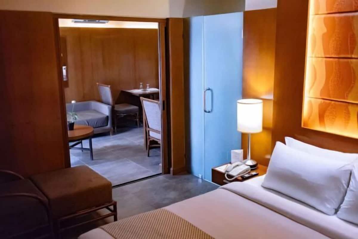 A hotel room with a bed, bedside table, lamp, and a view into a dining area with a table and chairs.