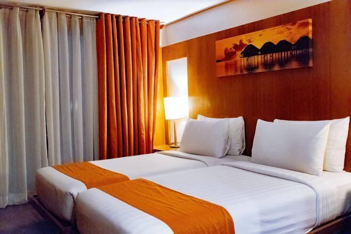 A double bed with white and orange bedding, a nightstand with a lamp, and a painting of a sunset on the wall.