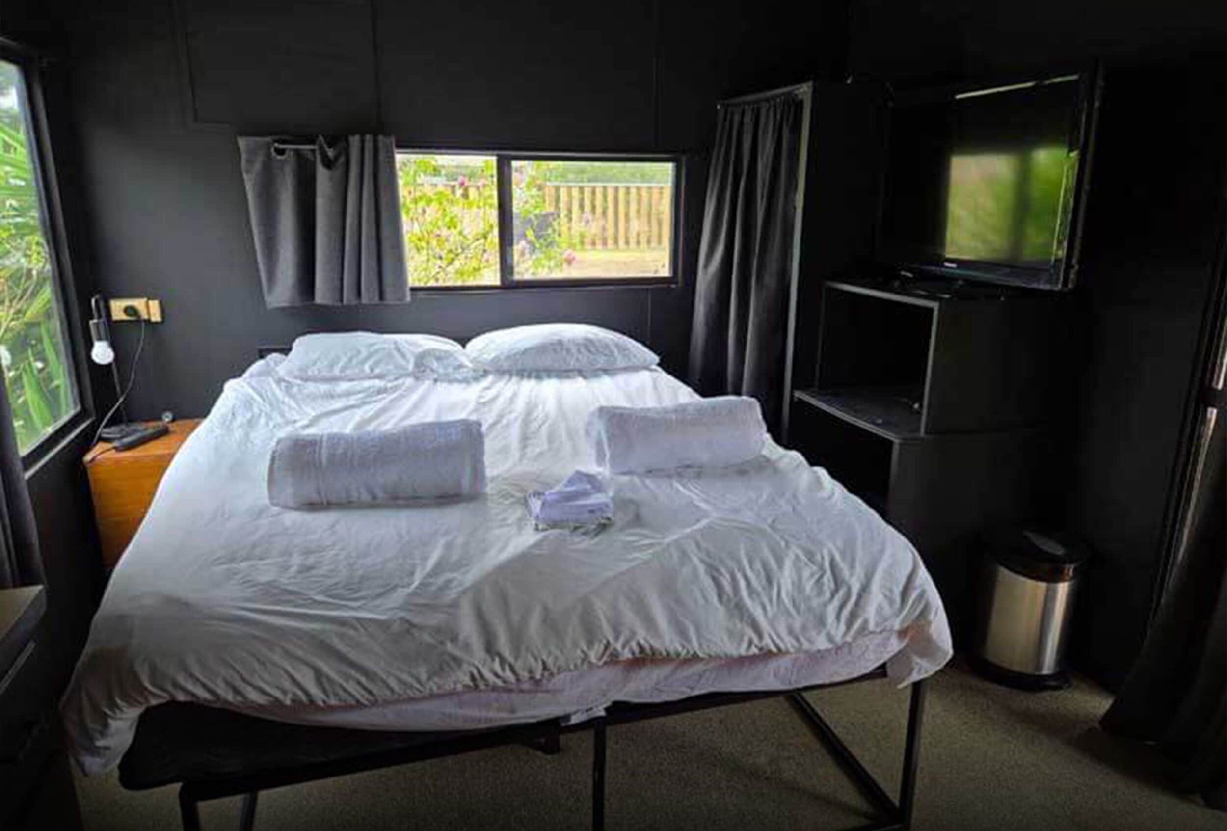 Un lit avec du linge de lit blanc, une table de chevet avec une lampe, un téléviseur fixé au mur et une fenêtre avec des rideaux.