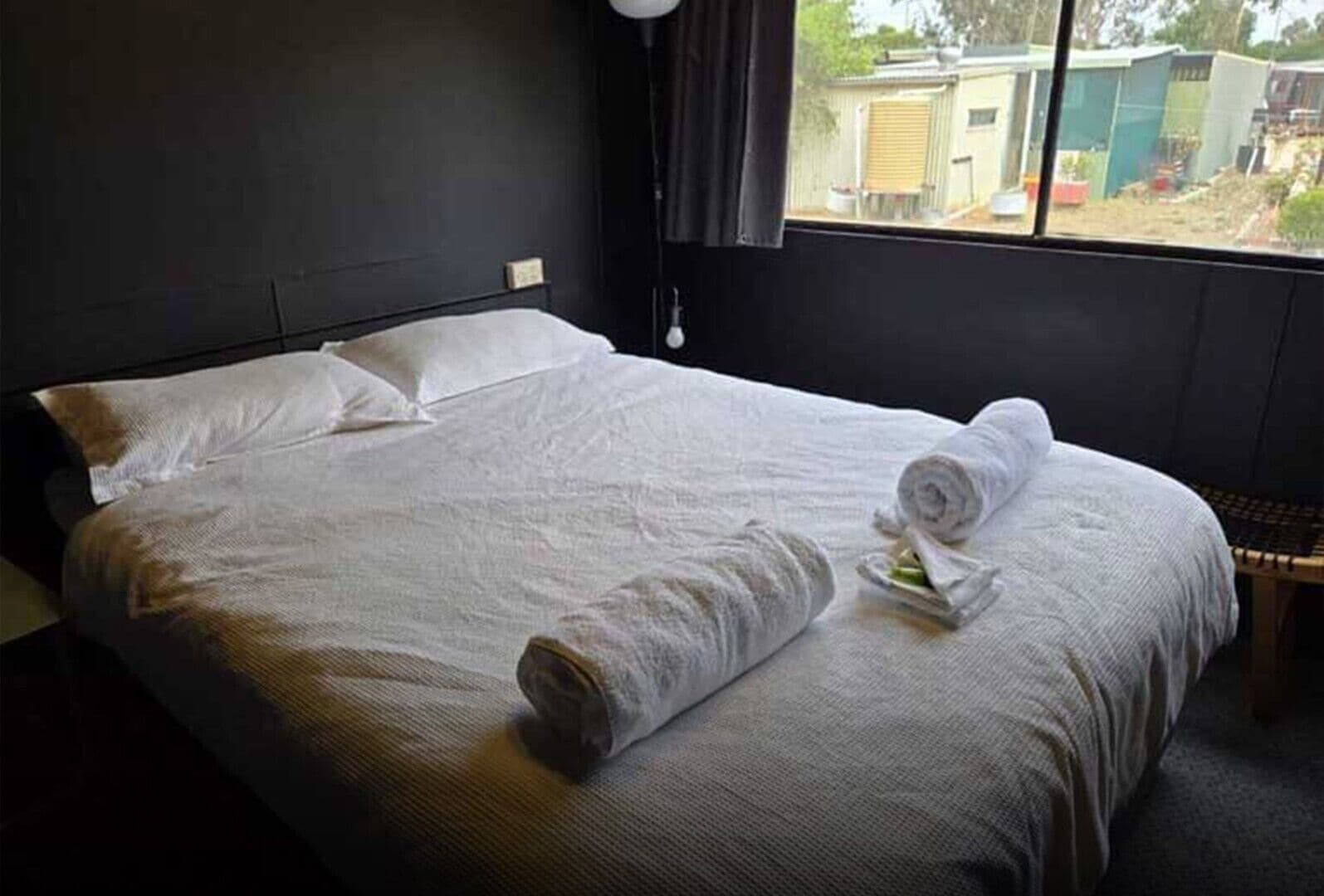 Un lit bien fait, avec du linge de lit blanc et une serviette enroulée, une fenêtre donnant sur un jardin, et un mur sombre.