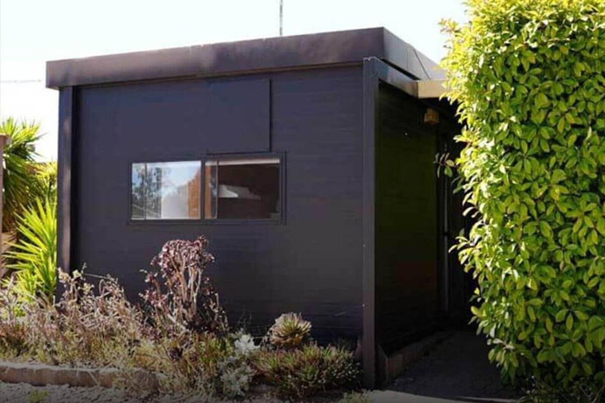 Un edificio moderno de color oscuro con una ventana pequeña y una puerta, rodeado de plantas y arbustos.
