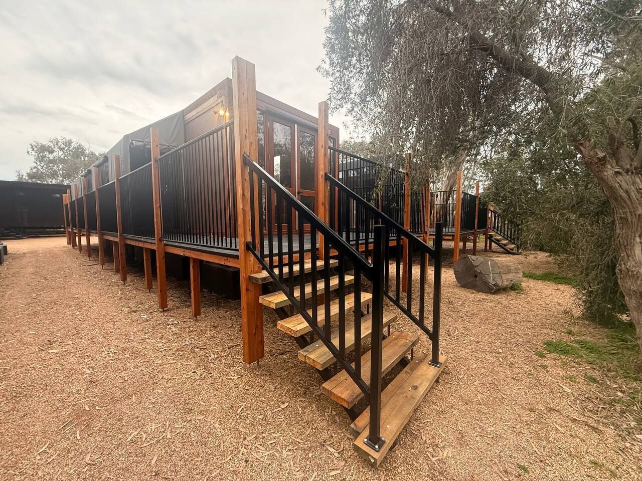 A wooden deck with a staircase leading to a covered area, surrounded by trees and a gravel ground.