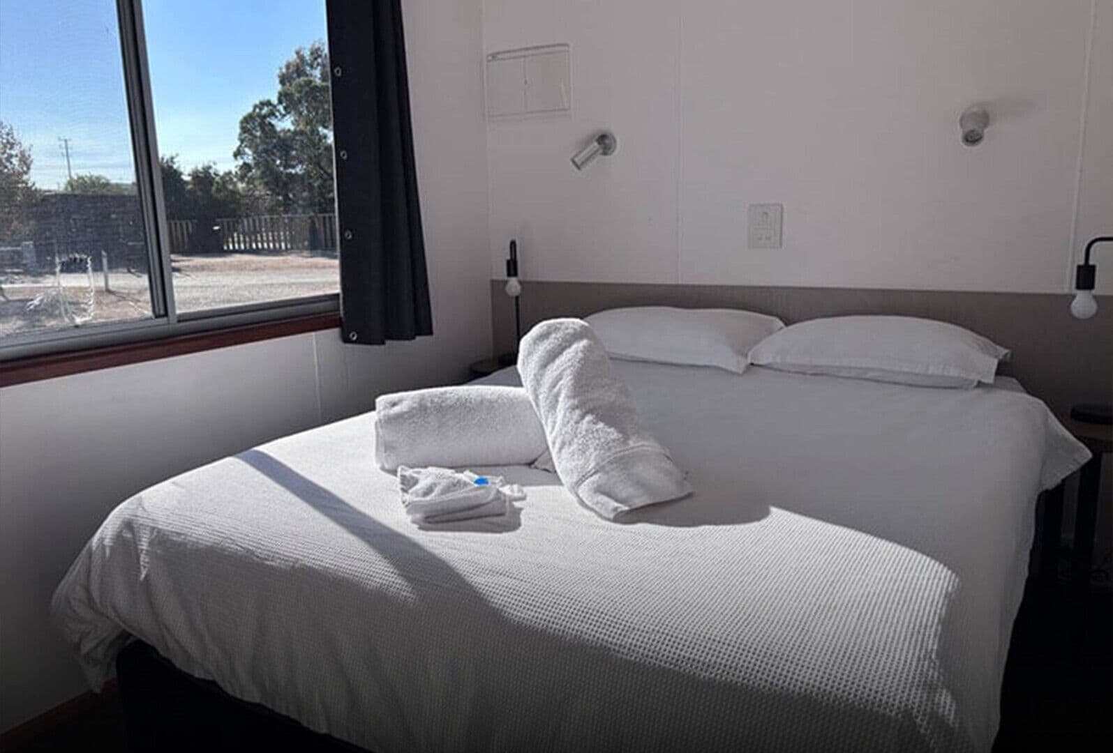 Una cama bien hecha con ropa blanca y toallas, una ventana con vista a un espacio exterior y una lámpara fijada a la pared.