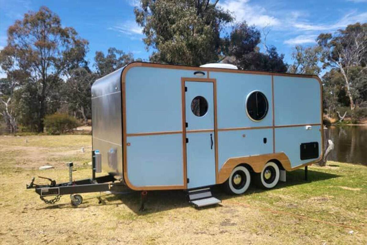 Una caravana recreativa azul y marrón aparcada sobre césped, con un cuerpo de agua y árboles al fondo.