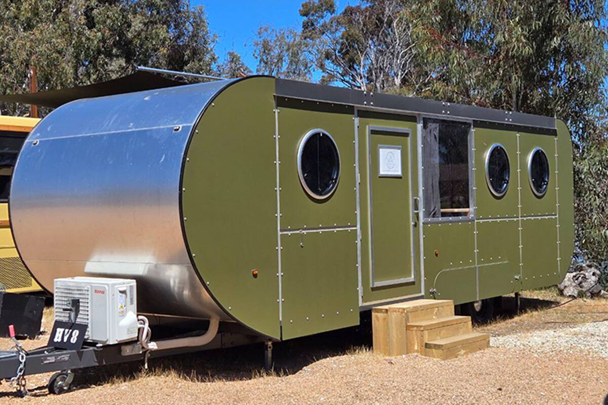 Una casa móvil cilíndrica verde, con un ventanal grande y una puerta pequeña, equipada con un aire acondicionado portátil.