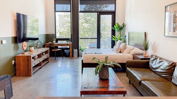 Una sala de estar moderna con una cama grande, un sofá, una mesa de centro de madera y un escritorio con una silla. Amplias ventanas ofrecen luz natural y vistas al exterior.