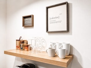 A wooden shelf with jars, glasses, and white mugs against a white wall.