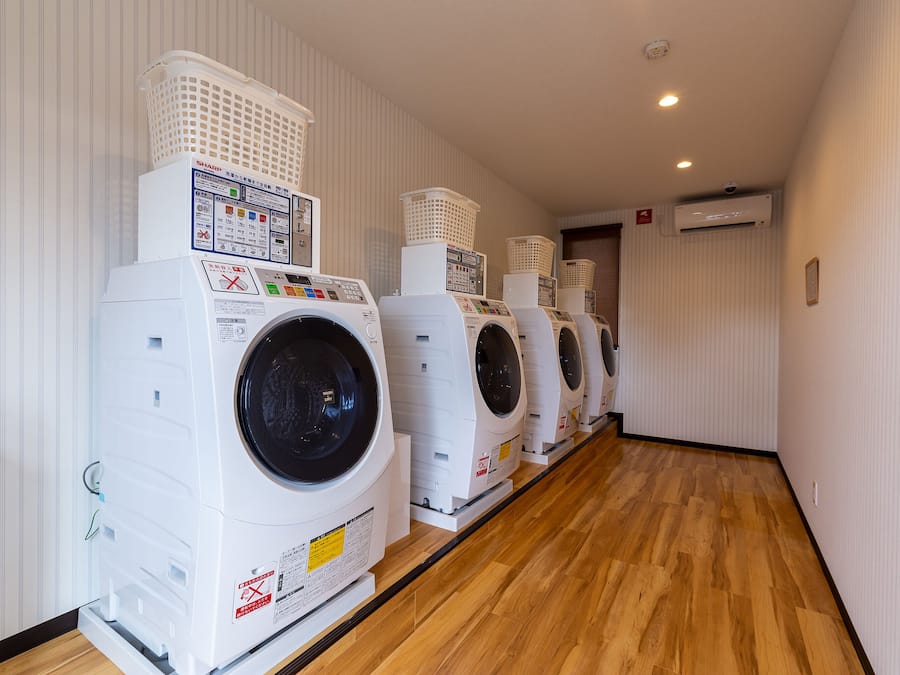 A row of white front-loading washing machines with baskets on top, in a hallway with wooden flooring and white walls.