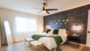 A modern bedroom with a brick wall, a bed with a green blanket, a wooden bench, and a ceiling fan. - Monte Horizon Oasis by HomeSlice Stays (Phoenix)