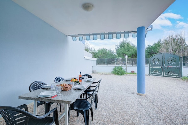 An outdoor dining area with a table set for four, surrounded by black chairs, under a covered section with a blue pillar.