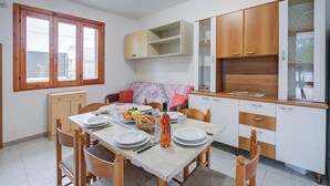 Uma área de refeições com uma mesa posta para quatro pessoas, cadeiras de madeira, um armário com prateleiras abertas e uma janela com molduras de madeira.