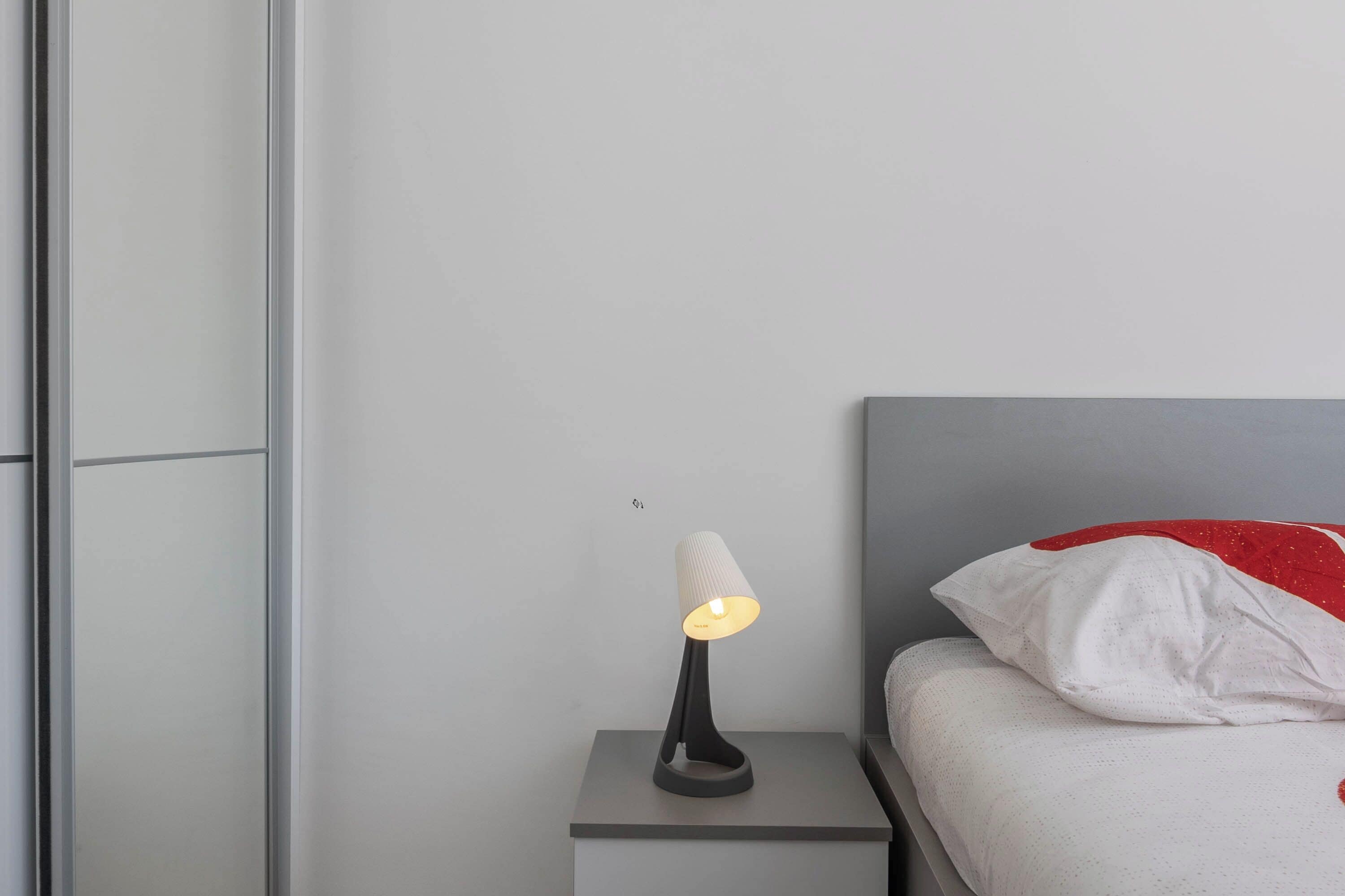 A modern bedroom with a bedside table, a lamp, a bed with a red pillow, and a grey headboard.