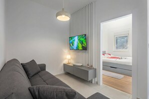 A modern living room with a grey sofa, a flat-screen TV mounted on a wall, a bedside table with a lamp, and a bed with a white headboard. - Seafront Villa Andrijana (Krsan)