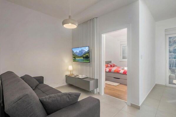 A modern living room with a grey sofa, a flat-screen TV mounted on a wall, a bedside table with a lamp, and a view into a bedroom with a bed and a window. - Seafront Villa Andrijana (Krsan)