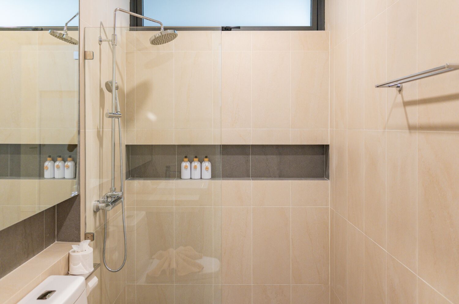 Une salle de bain moderne avec des carreaux de couleur beige, une douche en verre, un porte-serviettes et une étagère contenant des articles de toilette.