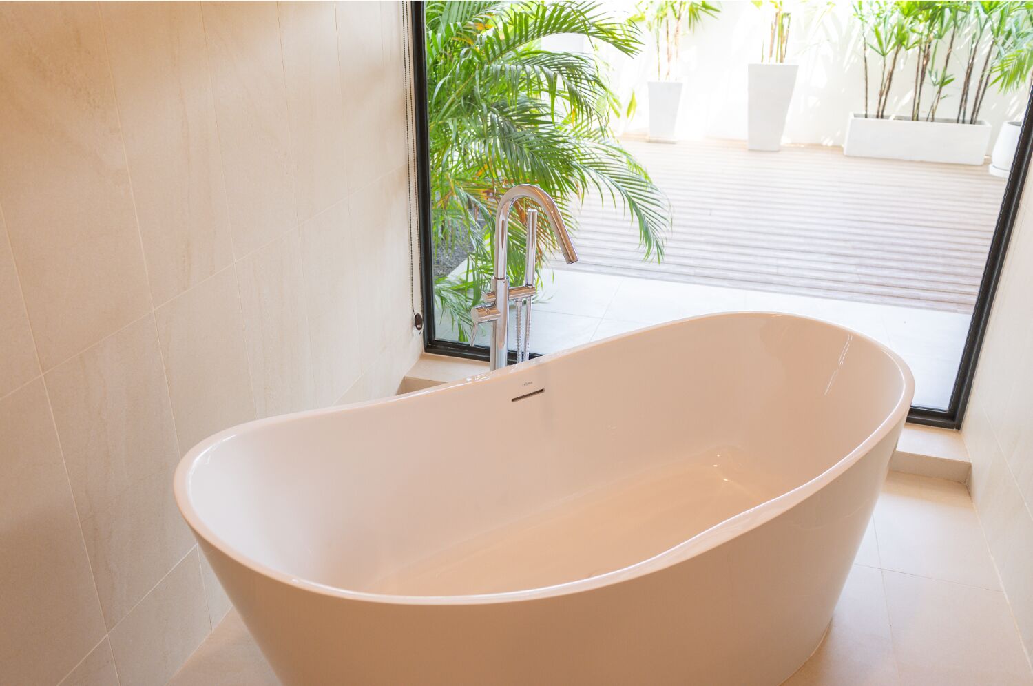 A modern bathroom with a freestanding bathtub, a window with a view of potted plants, and a tiled floor.