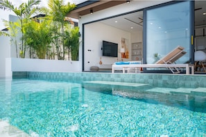 Un área de piscina exterior moderna con agua cristalina, rodeada de abundante vegetación y una casa contemporánea con amplias ventanas.