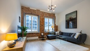 Un salon moderne avec un mur en briques, de grandes fenêtres, un canapé gris, un tapis blanc et un lustre.