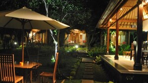 An outdoor dining area with wooden tables and chairs, a large umbrella, and a stone pathway leading to a building with a covered porch. - Kencana Garden Lembongan (Lembongan Island)